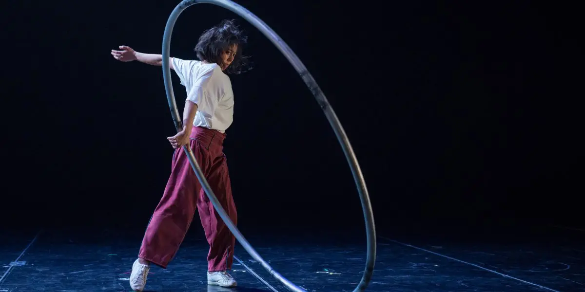 Miho Inaba, une artiste en pantalon rouge et chemise blanche, fait tourner un grand cerceau de métal sur une scène sombre, capturée en plein mouvement avec une pose dramatique.