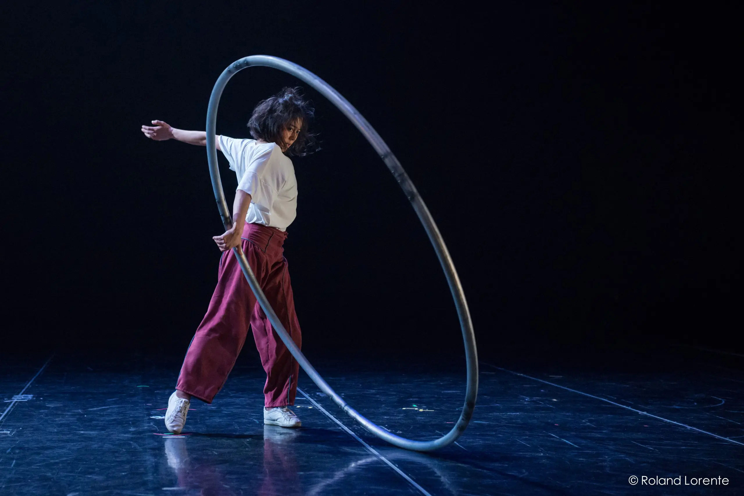 Miho Inaba, une artiste en pantalon rouge et chemise blanche, fait tourner un grand cerceau de métal sur une scène sombre, capturée en plein mouvement avec une pose dramatique.