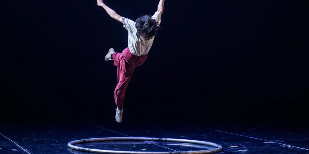 Miho Inaba, vêtue d'un pantalon rouge et d'une chemise blanche, saute gracieusement au-dessus d'un cerceau circulaire sur une scène sombre, les bras tendus et les jambes pliées, capturée en plein vol sous un éclairage dramatique.