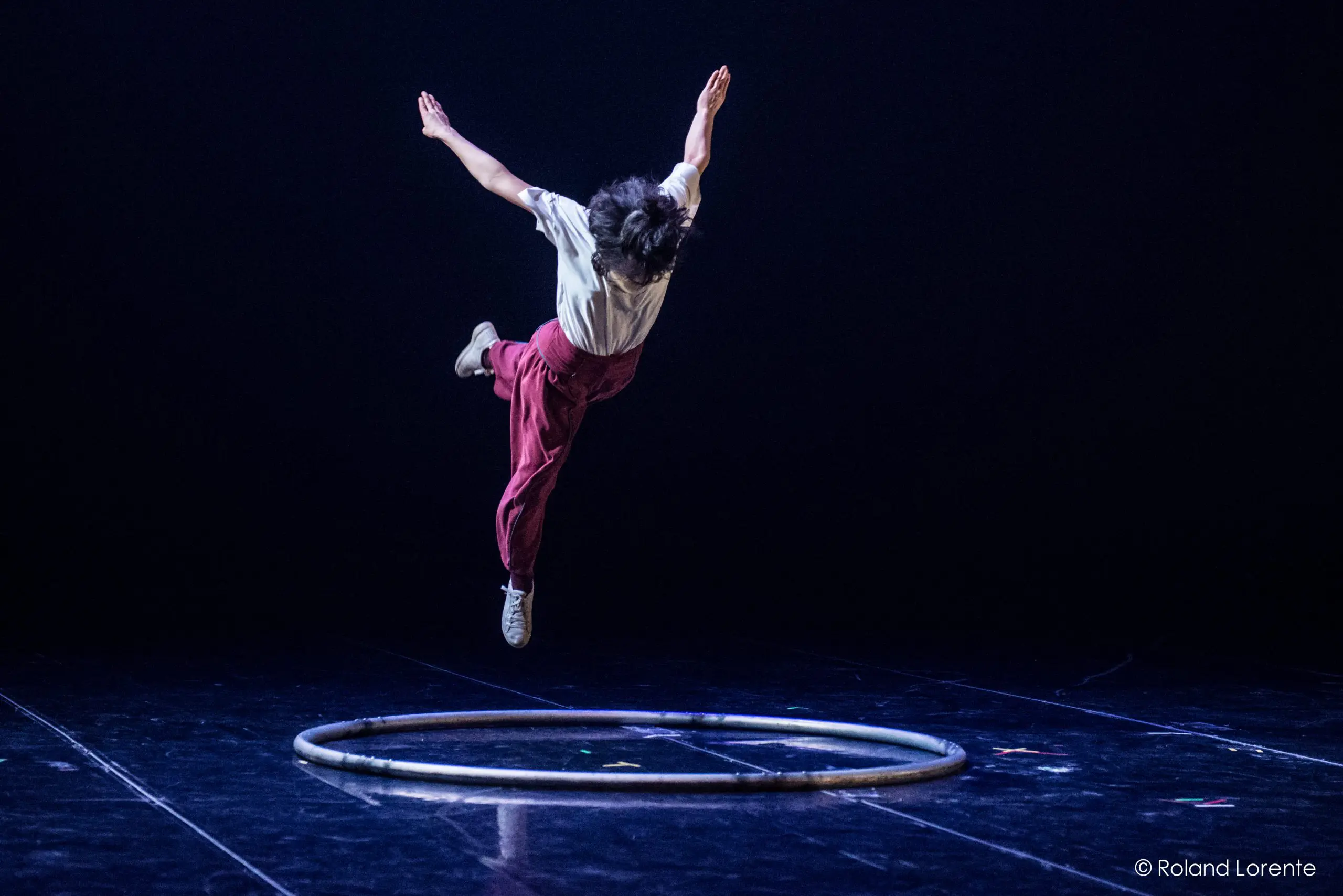 Miho Inaba, vêtue d'un pantalon rouge et d'une chemise blanche, saute gracieusement au-dessus d'un cerceau circulaire sur une scène sombre, les bras tendus et les jambes pliées, capturée en plein vol sous un éclairage dramatique.