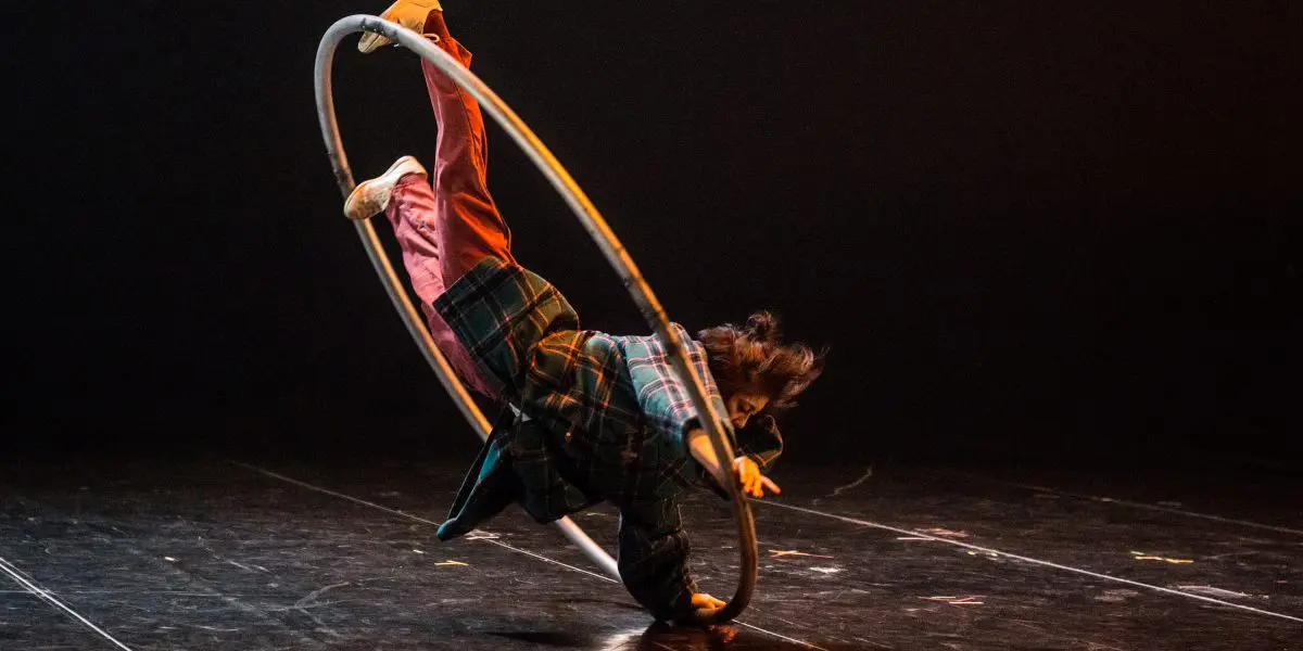 Miho Inaba, vêtue d'une chemise à carreaux et d'un pantalon rouge, se tient en équilibre dynamique à l'intérieur d'un grand cerceau métallique sur une scène sombre, une jambe tendue vers le haut et les bras en extension.