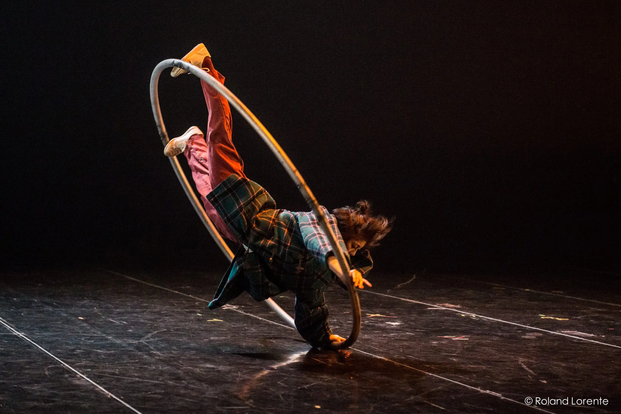 Miho Inaba, vêtue d'une chemise à carreaux et d'un pantalon rouge, se tient en équilibre dynamique à l'intérieur d'un grand cerceau métallique sur une scène sombre, une jambe tendue vers le haut et les bras en extension.