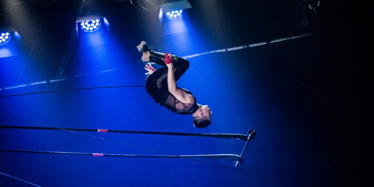 Nicolas Allard, acrobate, effectue un saut périlleux au-dessus d'un trapèze, éclairé par des lumières de scène bleues sur un fond sombre, lors d'un numéro de cirque.