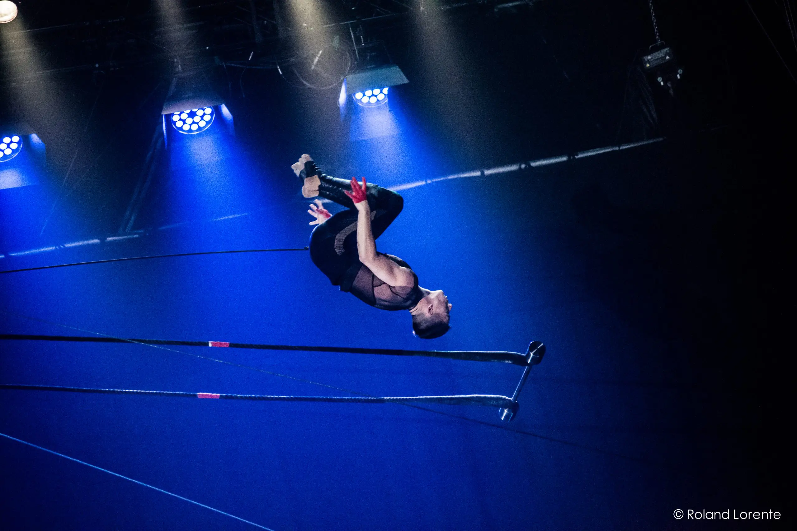 Nicolas Allard, acrobate, effectue un saut périlleux au-dessus d'un trapèze, éclairé par des lumières de scène bleues sur un fond sombre, lors d'un numéro de cirque.