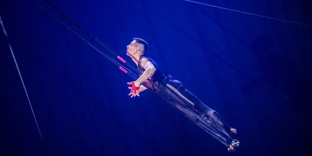 Un acrobate masculin, Nicolas Allard, est suspendu à des sangles noires, arquant le dos et regardant vers le haut sur un fond de scène éclairé en bleu.