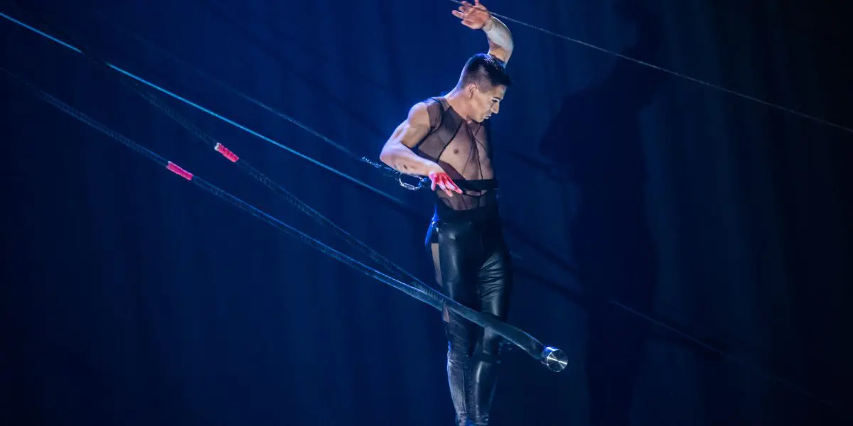 Un artiste aérien, Nicolas Allard, en pantalon noir, se tient en équilibre sur un appareil suspendu avec des cordes, baigné dans un éclairage de scène bleu et projetant une ombre sur la toile de fond.