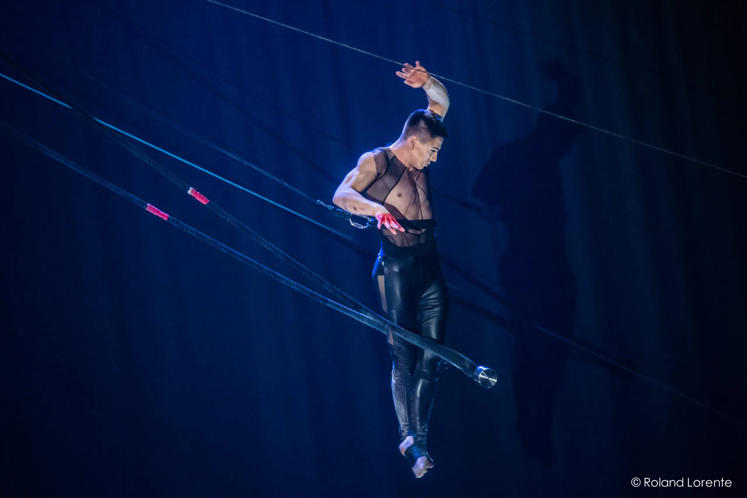 Un artiste aérien, Nicolas Allard, en pantalon noir, se tient en équilibre sur un appareil suspendu avec des cordes, baigné dans un éclairage de scène bleu et projetant une ombre sur la toile de fond.