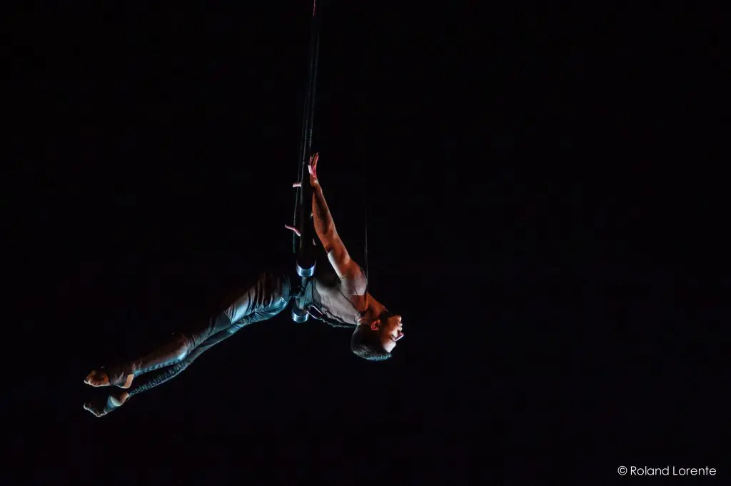 Un artiste, inspiré par l'art de Nicolas Allard, est suspendu dans les airs par des harnais et des cordes, arquant son dos de façon spectaculaire sur un fond sombre, éclairé par la lumière de la scène.