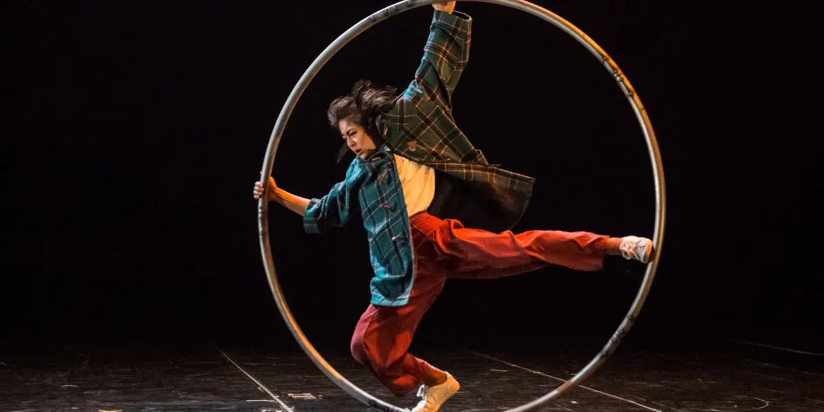 Miho Inaba, vêtue de vêtements amples, se tient habilement en équilibre à l'intérieur d'un grand cerceau métallique sur une scène sombre, exécutant un numéro de cirque ou d'acrobatie impressionnant.