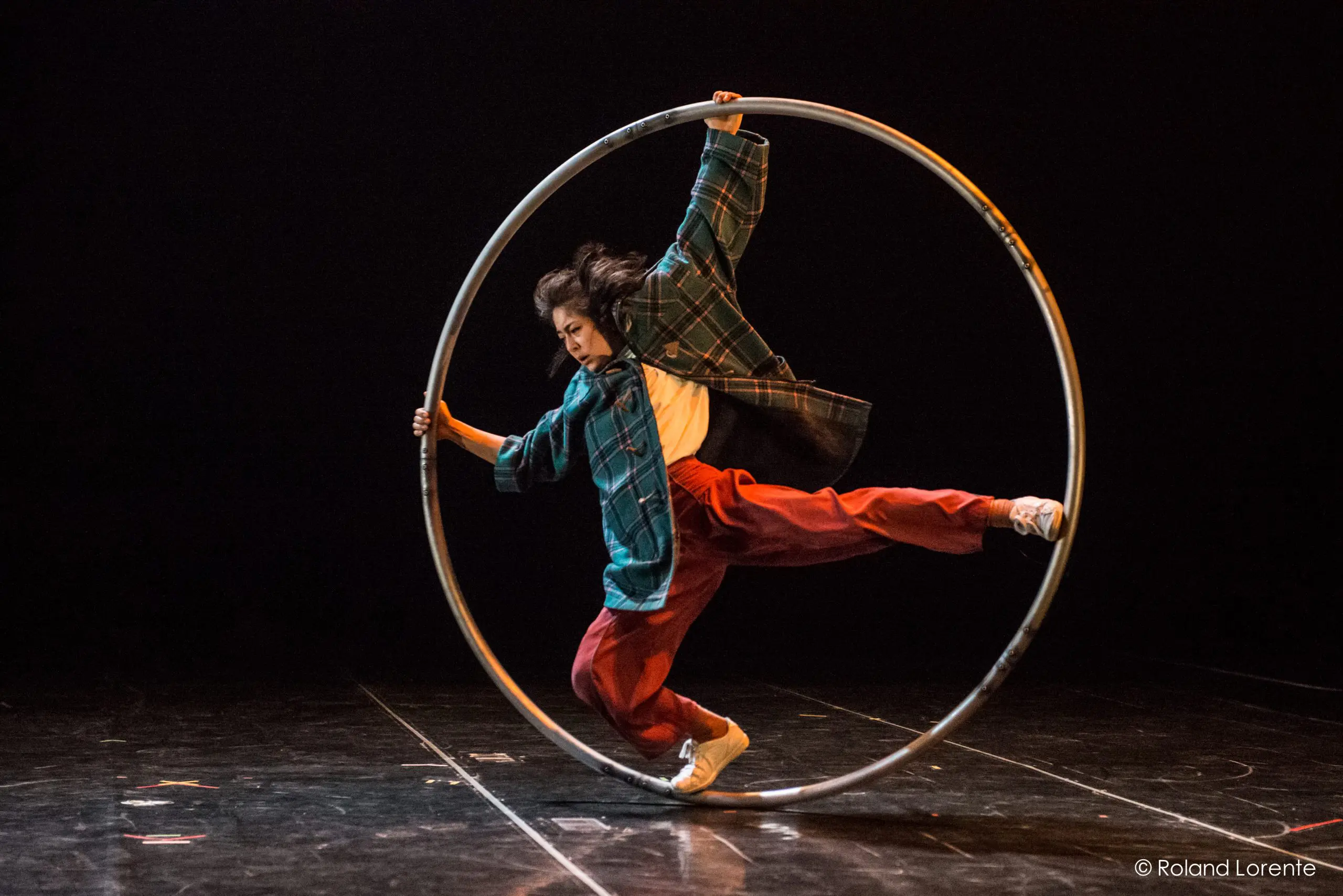 Miho Inaba, vêtue de vêtements amples, se tient habilement en équilibre à l'intérieur d'un grand cerceau métallique sur une scène sombre, exécutant un numéro de cirque ou d'acrobatie impressionnant.