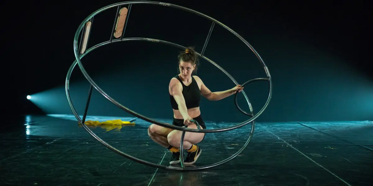 Clara Prezzavento, vêtue d'une tenue de sport, est accroupie à l'intérieur d'une grande roue métallique sur une scène sombre éclairée par un éclairage dramatique, avec un tissu jaune posé à proximité.