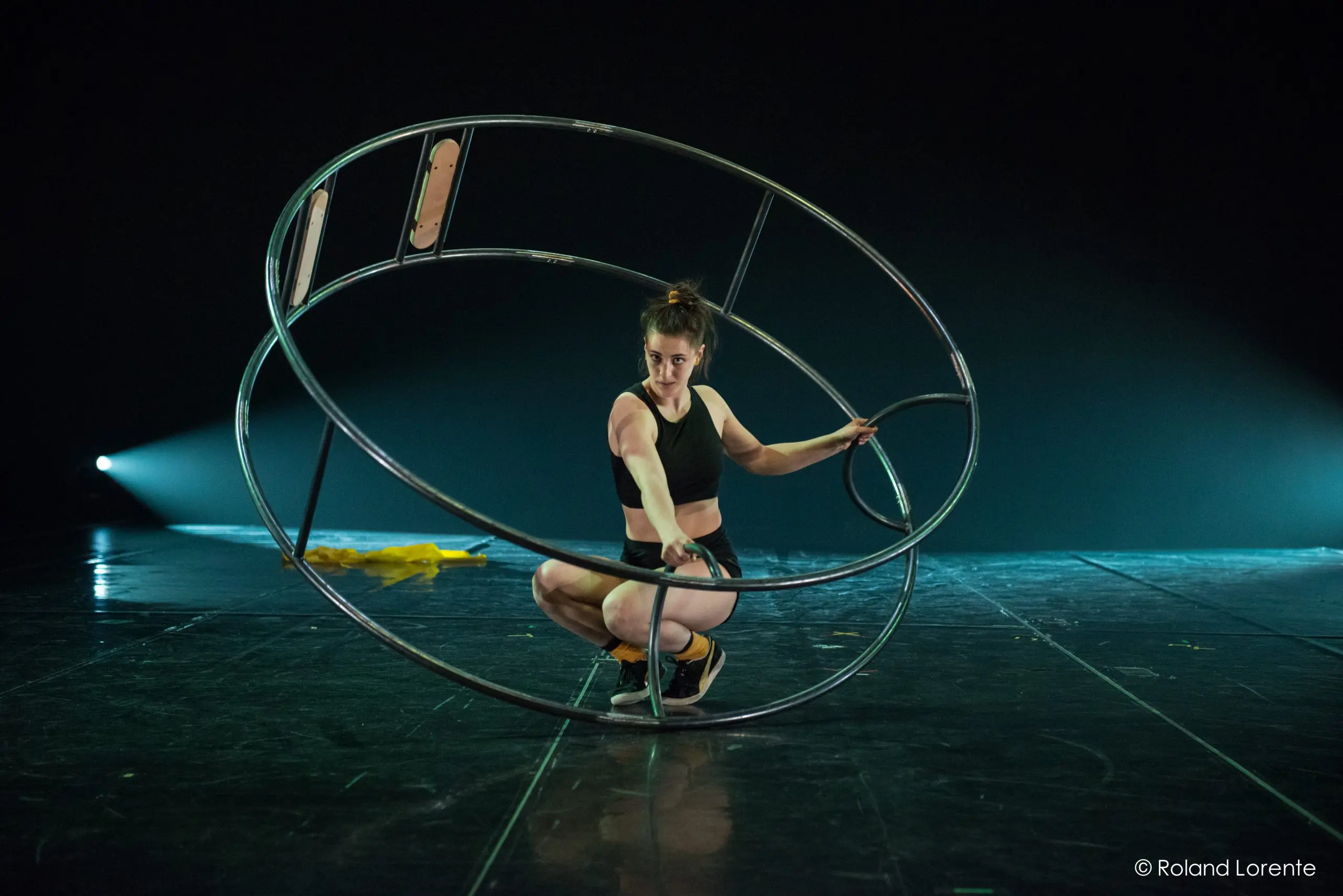 Clara Prezzavento, vêtue d'une tenue de sport, est accroupie à l'intérieur d'une grande roue métallique sur une scène sombre éclairée par un éclairage dramatique, avec un tissu jaune posé à proximité.