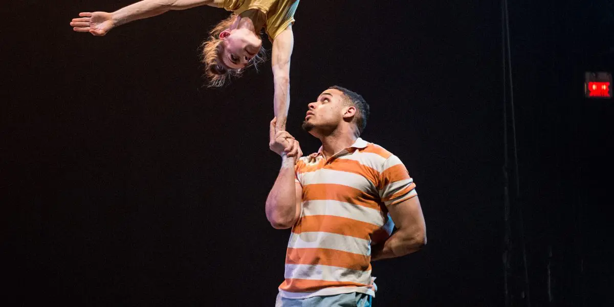 Un interprète masculin, Michael Patterson, en chemise rayée, tient Joëlle Ziorjen la tête en bas sur une main ; elle étire son corps gracieusement, les jambes étendues, sur un fond de scène sombre.