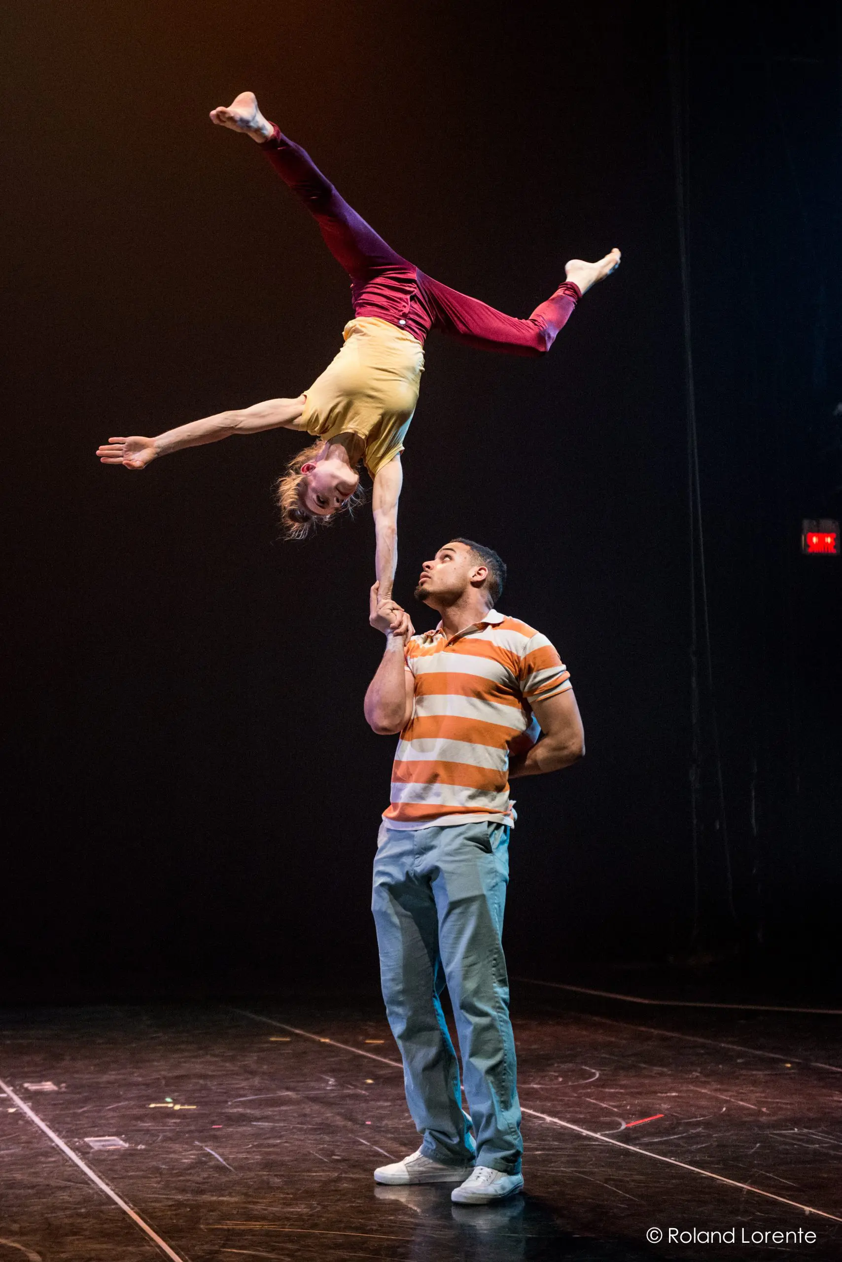Un interprète masculin, Michael Patterson, en chemise rayée, tient Joëlle Ziorjen la tête en bas sur une main ; elle étire son corps gracieusement, les jambes étendues, sur un fond de scène sombre.