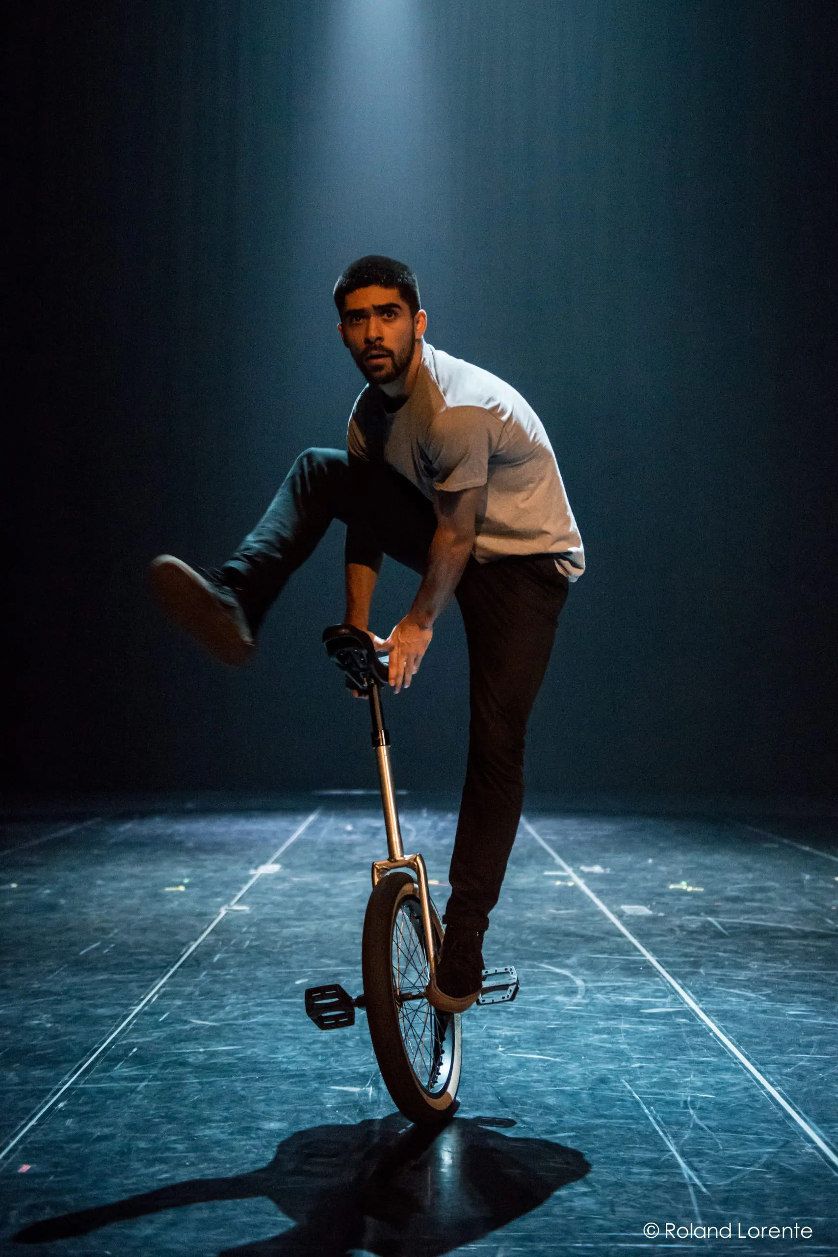 Ruben Ingwersen exécute un tour sur un monocycle sous un éclairage dramatique, en équilibre avec une jambe tendue tout en traversant une scène sombre et éclairée par des projecteurs.