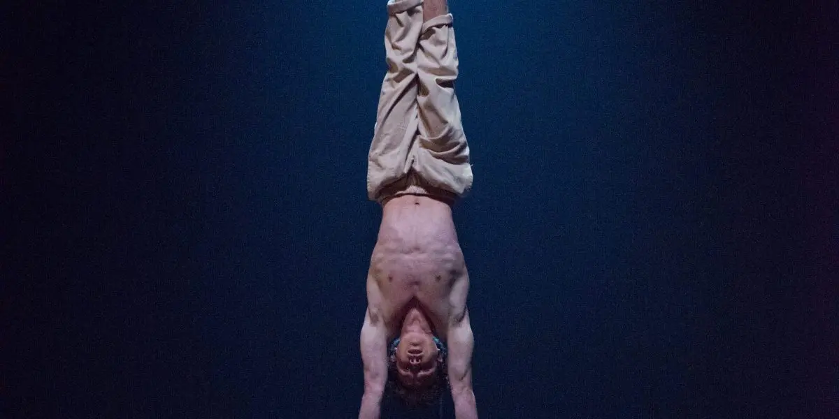 Un artiste torse nu, Jarrod Takle, se tient en équilibre sur des barres parallèles sous un projecteur sur une scène sombre, vêtu d'un pantalon beige ample.