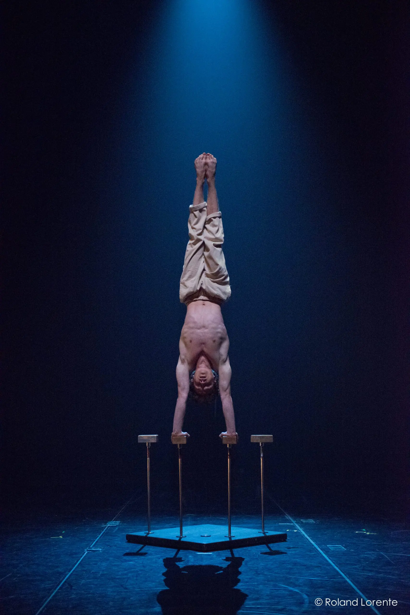Un artiste torse nu, Jarrod Takle, se tient en équilibre sur des barres parallèles sous un projecteur sur une scène sombre, vêtu d'un pantalon beige ample.