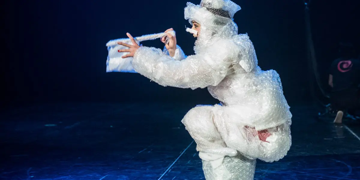 Une artiste vêtue de papier bulle et de bottes blanches est accroupie sur scène, tenant un sac à main en papier bulle assorti. Un éclairage dramatique met en valeur la scène sur un fond sombre dans cette œuvre saisissante d'Éline Guélat.