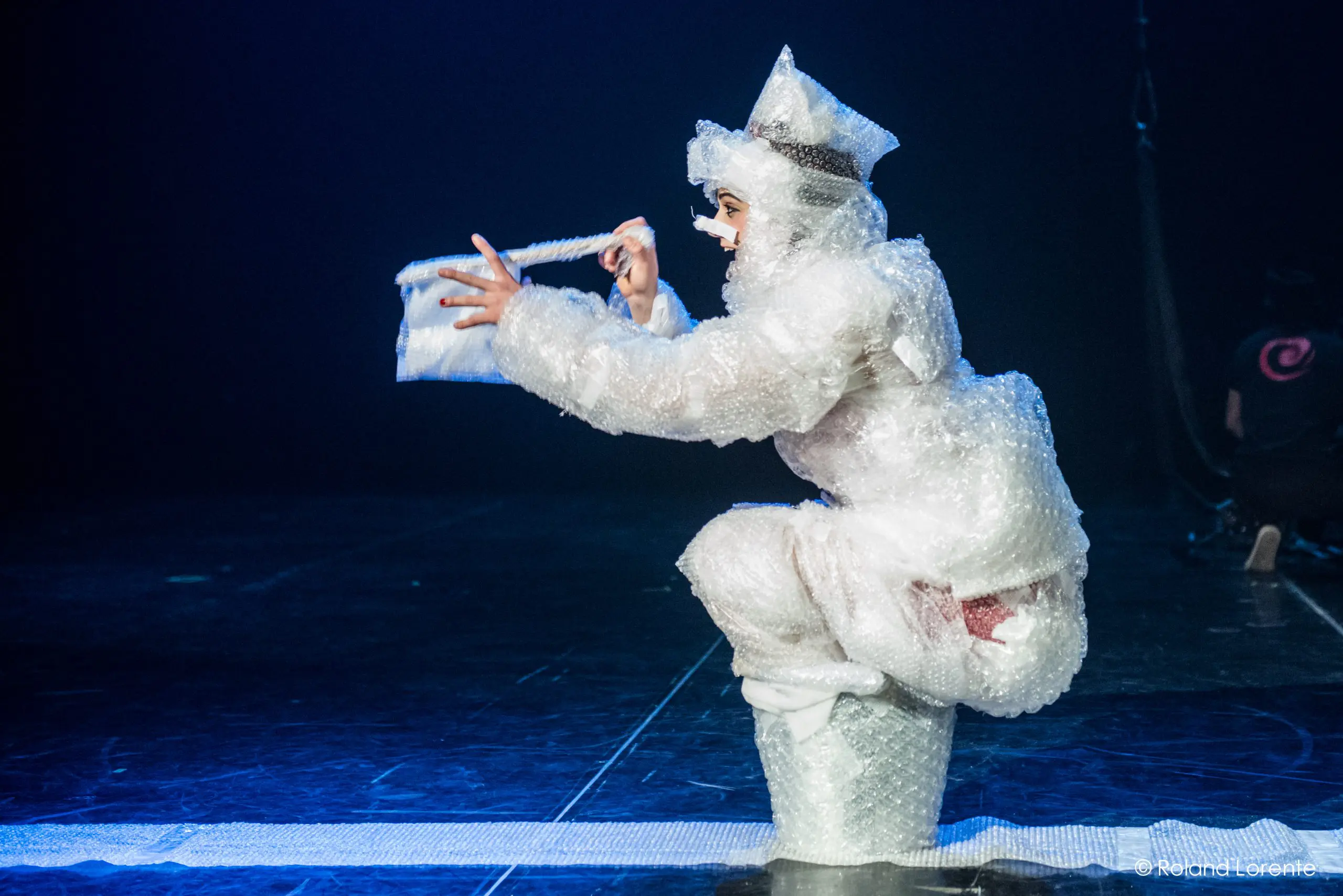 Une artiste vêtue de papier bulle et de bottes blanches est accroupie sur scène, tenant un sac à main en papier bulle assorti. Un éclairage dramatique met en valeur la scène sur un fond sombre dans cette œuvre saisissante d'Éline Guélat.