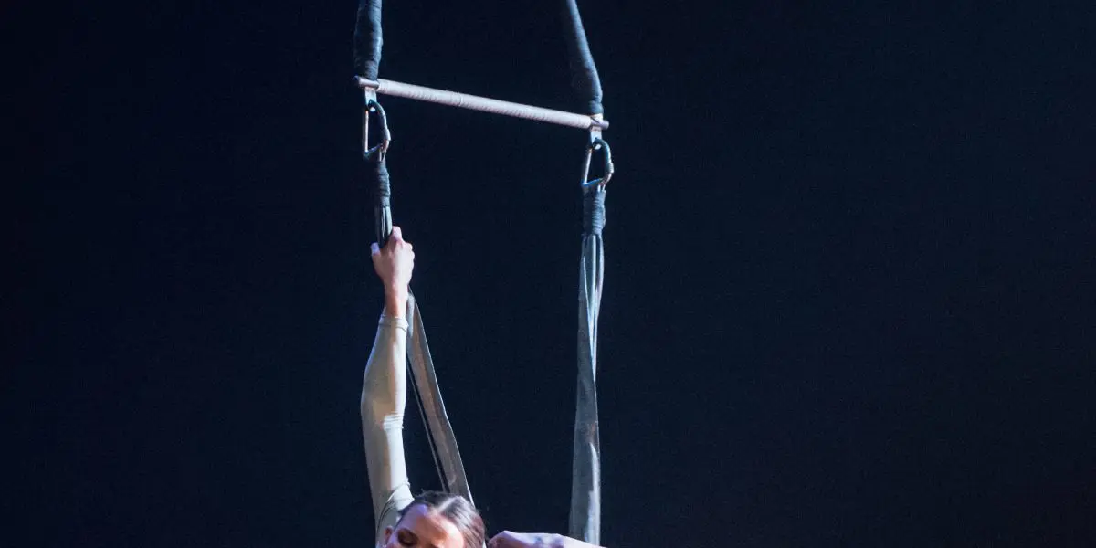 Une voltigeuse, Halle Baart, exécute une pose acrobatique suspendue à un trapèze, arquant gracieusement son dos et tenant un pied au-dessus de sa tête sur un fond sombre.
