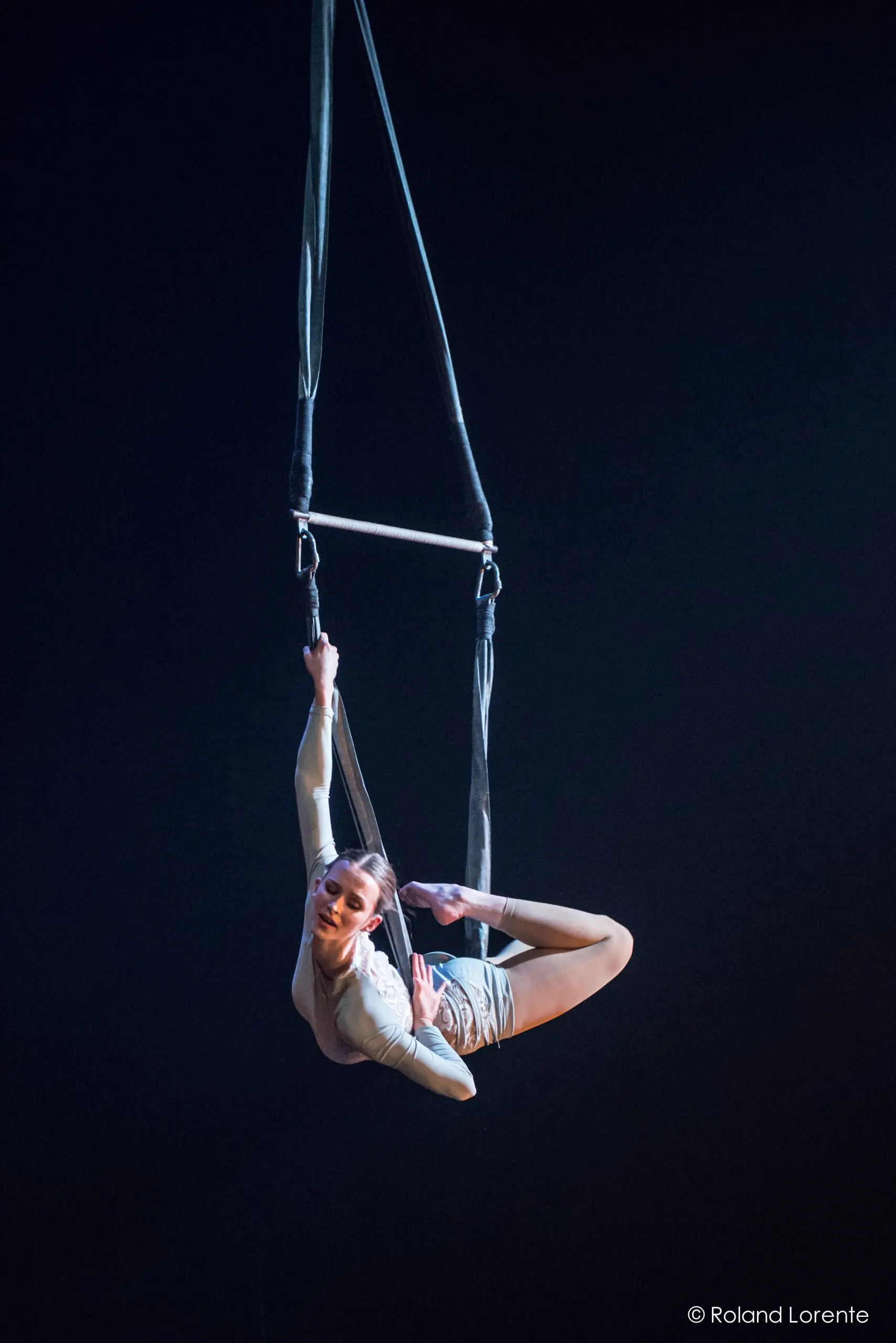 Une voltigeuse, Halle Baart, exécute une pose acrobatique suspendue à un trapèze, arquant gracieusement son dos et tenant un pied au-dessus de sa tête sur un fond sombre.