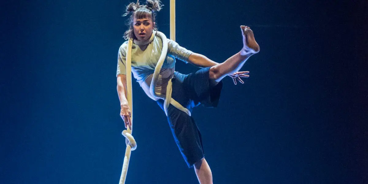 Un artiste de cirque, Sandy Tugwood, est suspendu à une longue corde aérienne blanche, une jambe pliée et un bras tendu. L'éclairage bleu de la scène met en valeur cette scène dramatique.