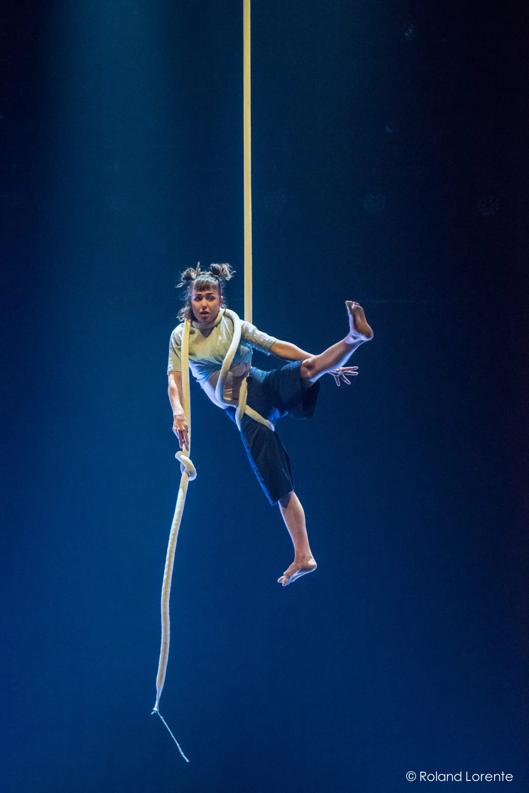 Un artiste de cirque, Sandy Tugwood, est suspendu à une longue corde aérienne blanche, une jambe pliée et un bras tendu. L'éclairage bleu de la scène met en valeur cette scène dramatique.