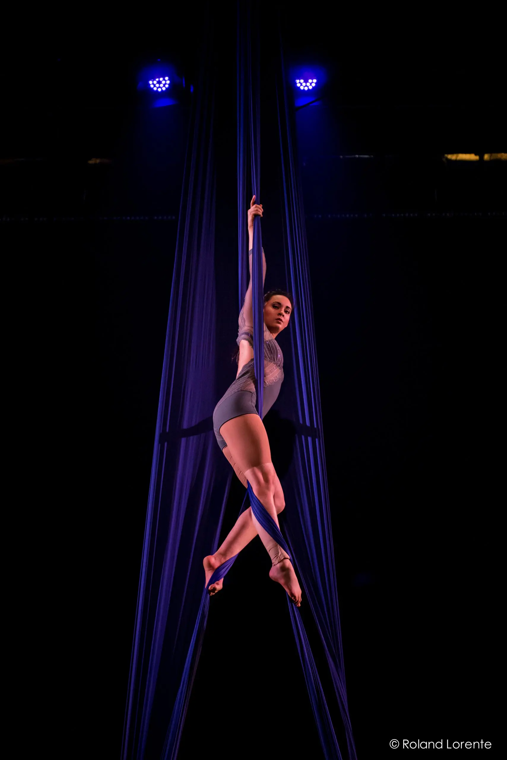Vanessa Aviles, une voltigeuse vêtue d'un justaucorps gris, se produit suspendue à des soies bleues sur un fond de scène sombre, éclairé par des projecteurs.