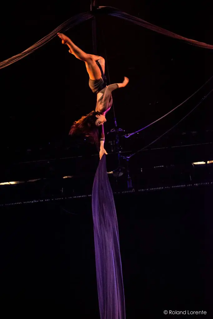 L'artiste aérienne Vanessa Aviles prend la pose à l'envers au-dessus de la scène, suspendue par des soies violettes sous un éclairage dramatique, avec l'obscurité en arrière-plan.