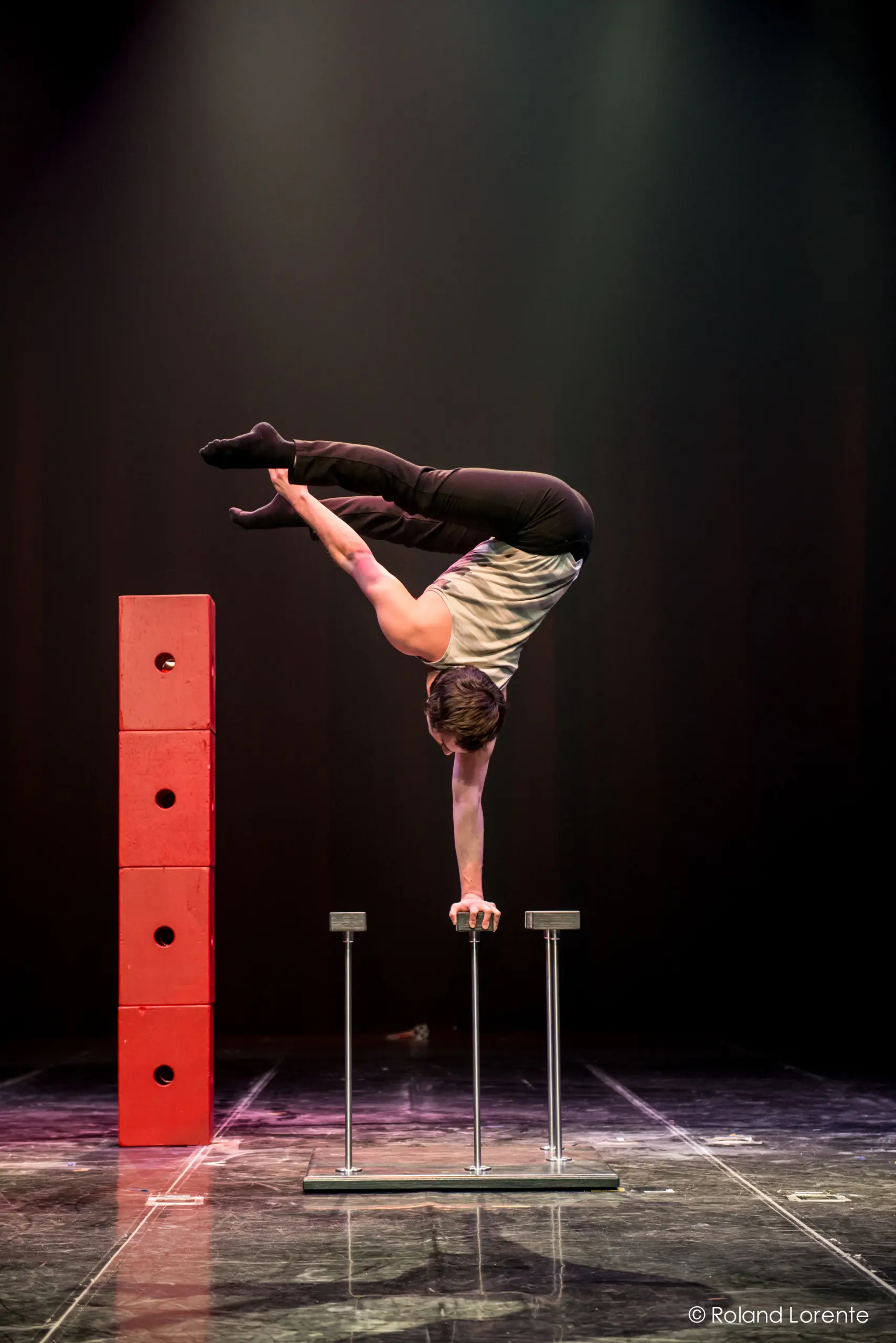 Le circassien Florian Jeannot se tient en équilibre sur une main au sommet d'une paire de cannes, les jambes tendues à l'horizontale, à côté d'une haute pile de blocs rouges sous un éclairage dramatique.