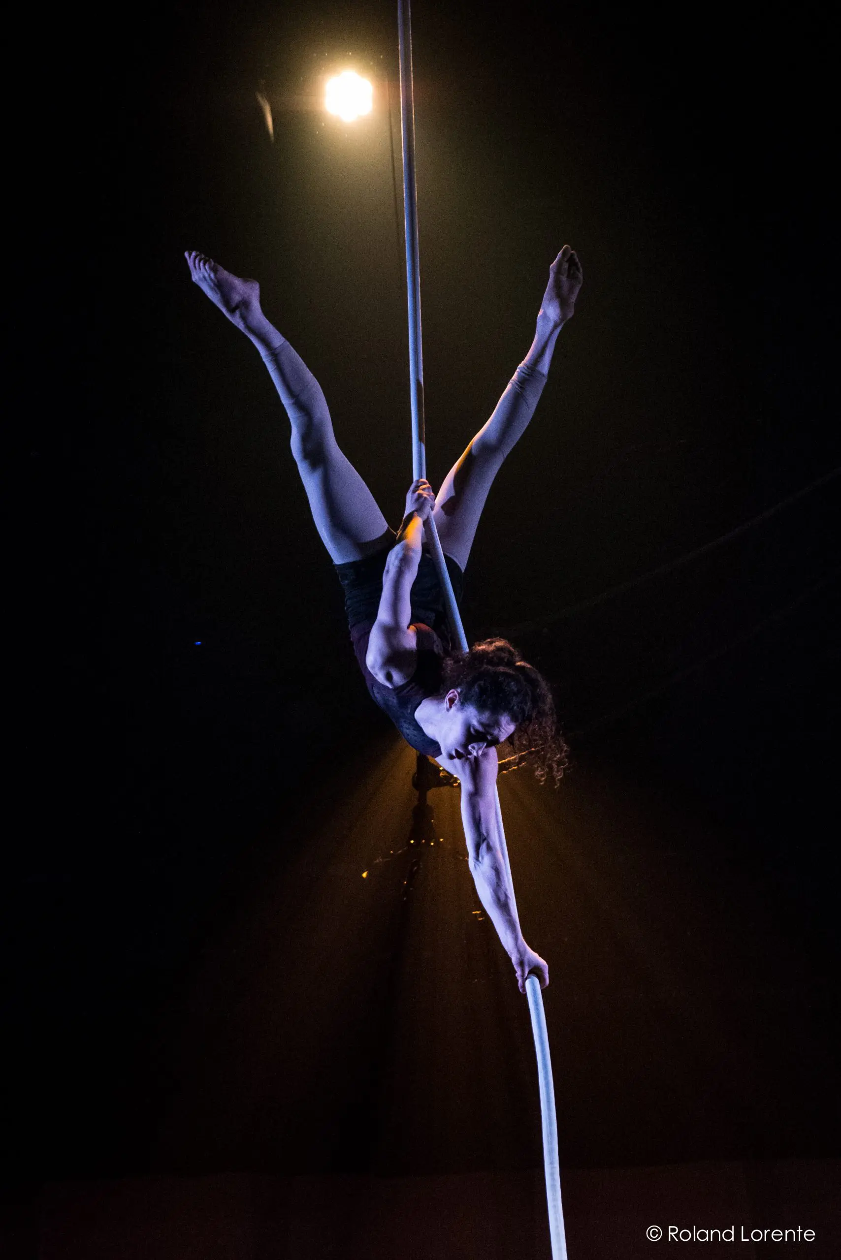 Une voltigeuse, Rosita Hendry, exécute une pose acrobatique sur une corde verticale, suspendue à l'envers sous un projecteur dans un théâtre sombre.