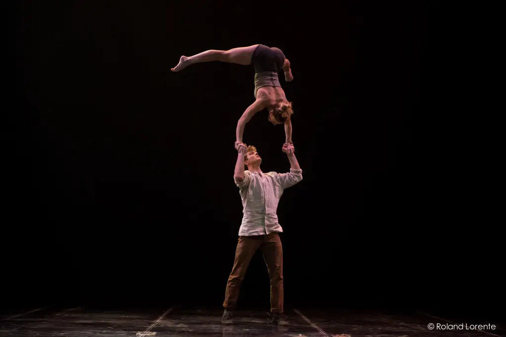 Andrei Anissimov et Emma Rogers sont captivés sur une scène sombre. Il la fait tenir en équilibre sur une main, la tête en bas, l'éclairage dramatique mettant en valeur leur force et leur équilibre. L'homme se tient fermement debout tandis que la femme tient une position de planche parfaite au-dessus de lui.