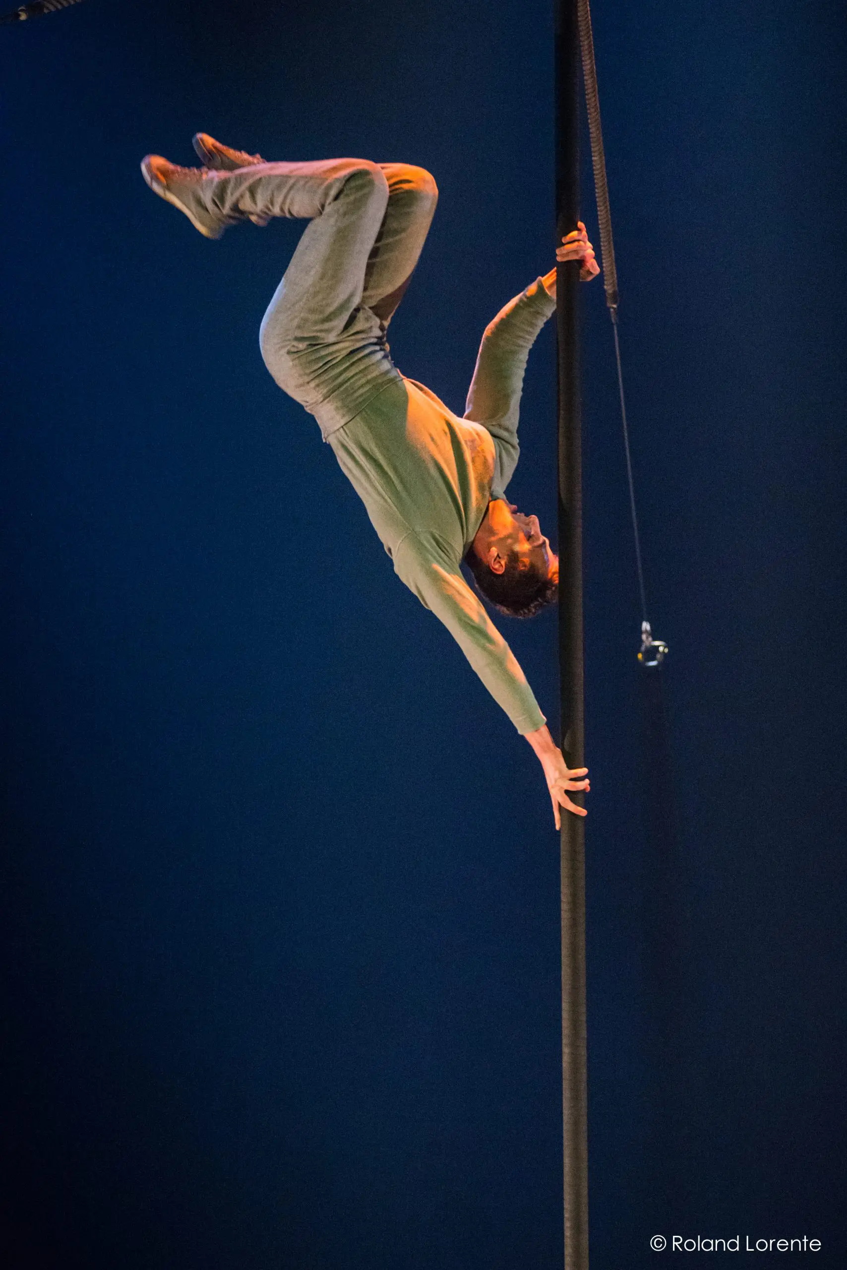 L'artiste Ron Oppenheimer, vêtu d'une chemise verte et d'un pantalon gris, est suspendu à l'envers à un poteau vertical, qu'il saisit d'une main et d'un pied, sur un fond bleu foncé.