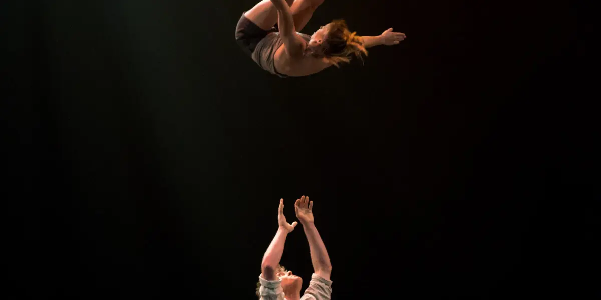 Un artiste, Andrei Anissimov, tend les bras tandis que l'acrobate Emma Rogers se balance au-dessus de lui sur une scène sombre, éclairée par des projecteurs spectaculaires.