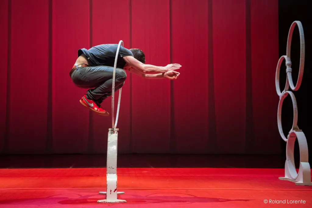 Un artiste vêtu d'une chemise bleue et de chaussures rouges saute dans un cerceau blanc sur une scène éclairée en rouge. Trois autres cerceaux empilés sont visibles sur le côté droit de l'image, rappelant les numéros de Quentin Gréco et d'Evan Tomlinson Weintraub.