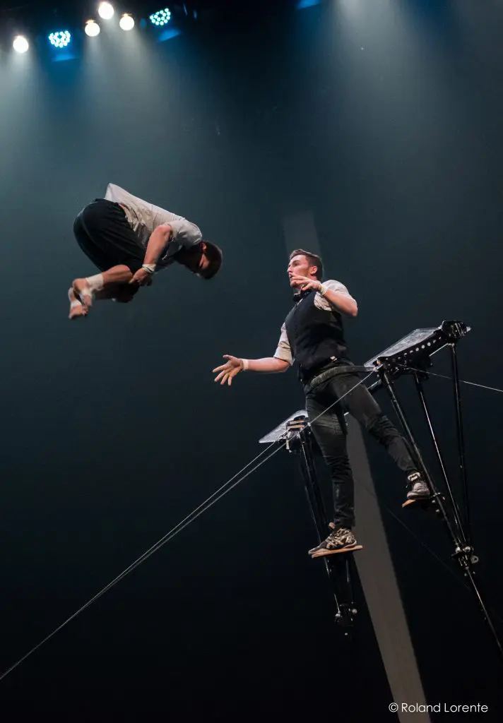 Un artiste fait des pirouettes en plein vol tandis que Maxime Blanckaert, debout sur une plate-forme surélevée, tend la main pour l'attraper lors d'un numéro de cirque sous les lumières de la scène.