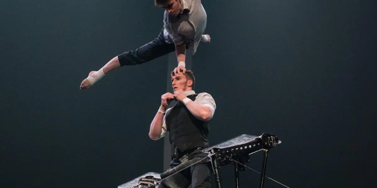 Un acrobate, Nathan Briscoe, se tient en équilibre sur des échasses tout en soutenant Maxime Blanckaert au-dessus de lui, en la tenant par la bouche. Elle pose gracieusement dans les airs, une jambe tendue, sous les lumières de la scène.