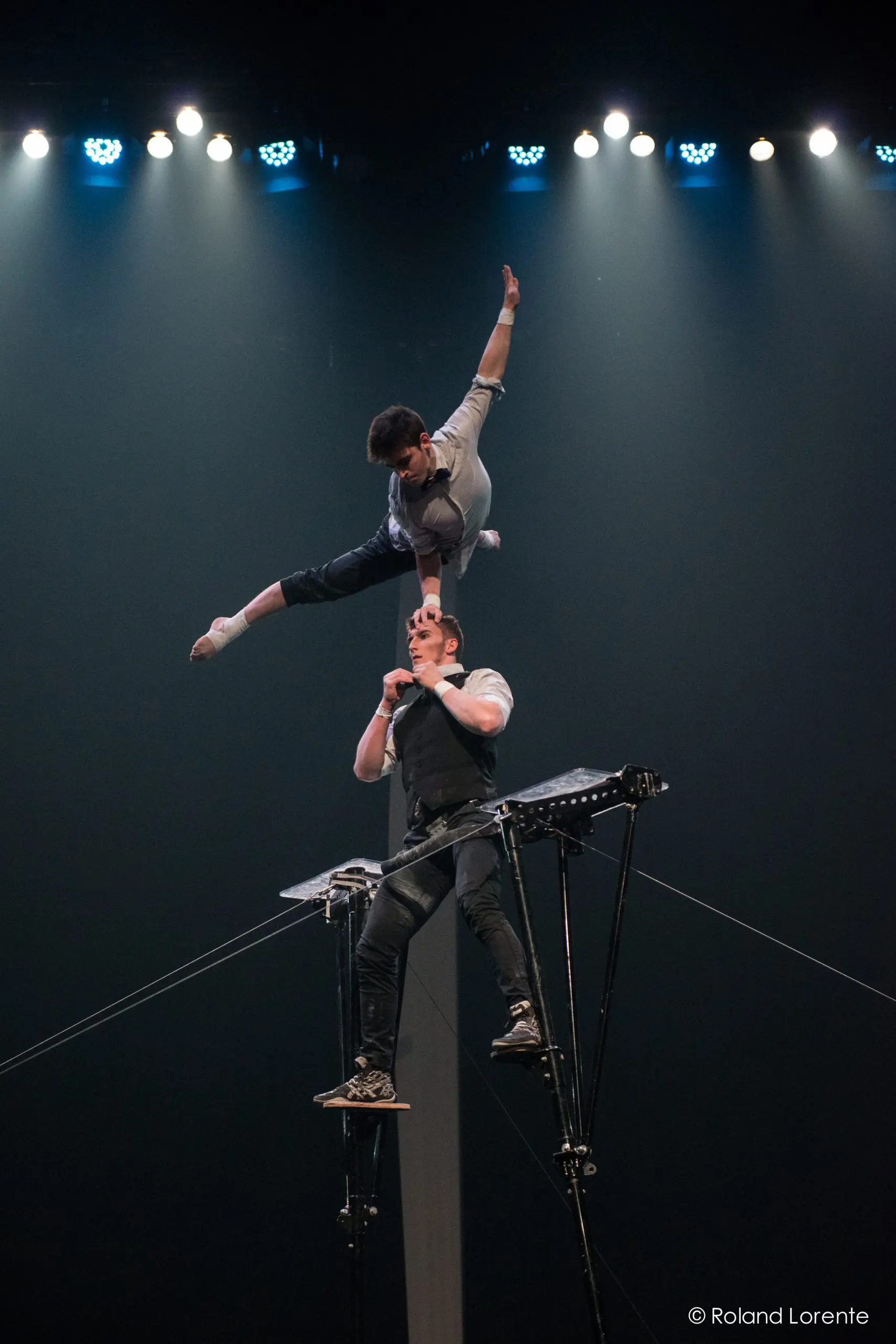 Un acrobate, Nathan Briscoe, se tient en équilibre sur des échasses tout en soutenant Maxime Blanckaert au-dessus de lui, en la tenant par la bouche. Elle pose gracieusement dans les airs, une jambe tendue, sous les lumières de la scène.