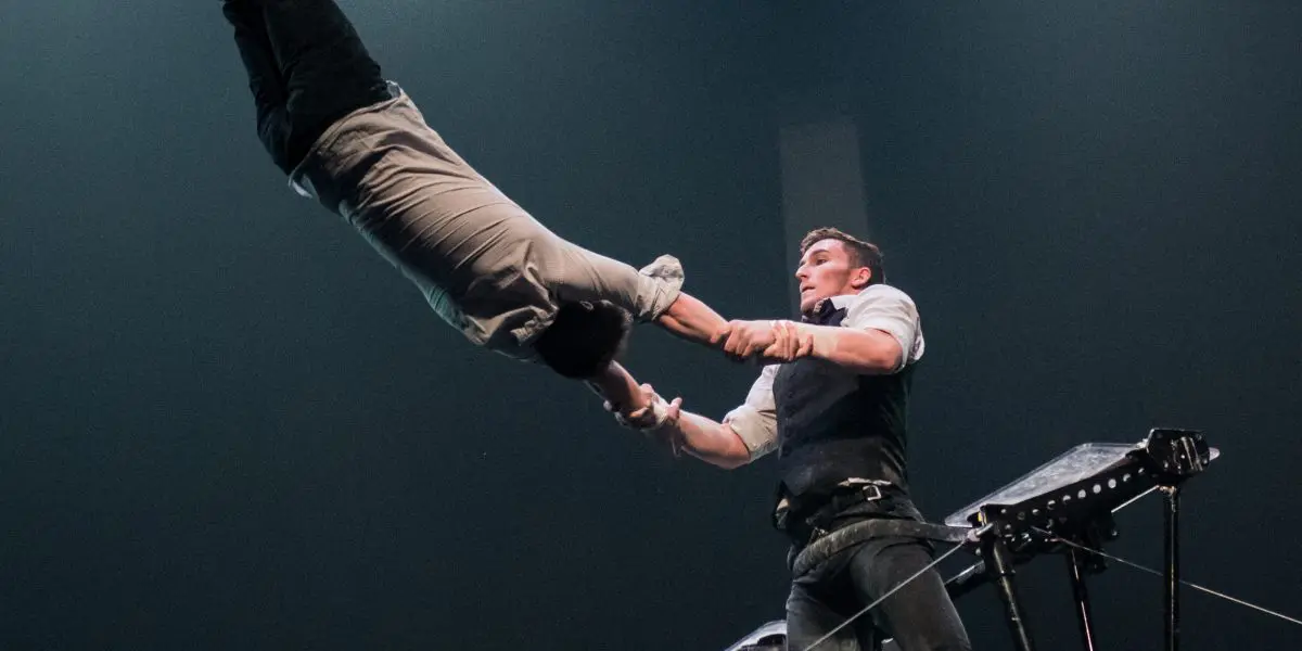 Maxime Blanckaert et Nathan Briscoe réalisent un audacieux numéro d'équilibre sur un fil de fer, l'un s'élançant dans les airs tandis que l'autre le soutient, le tout sous un éclairage scénique spectaculaire.