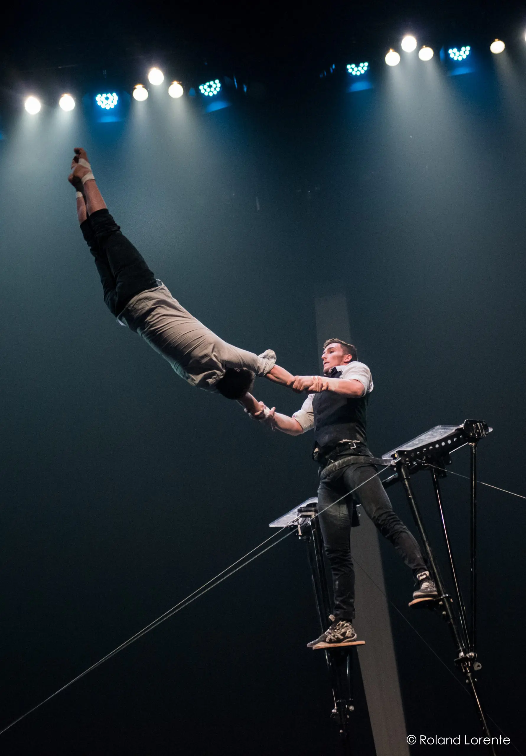Maxime Blanckaert et Nathan Briscoe réalisent un audacieux numéro d'équilibre sur un fil de fer, l'un s'élançant dans les airs tandis que l'autre le soutient, le tout sous un éclairage scénique spectaculaire.