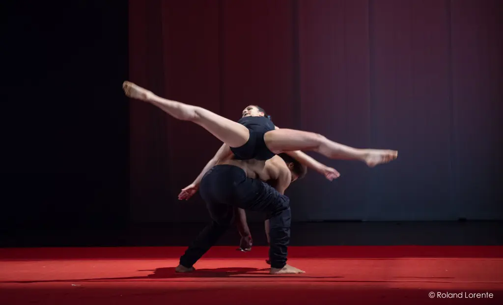 Marlon Archer et Jenay Espinosa sont captivés par l'envol acrobatique d'un danseur en noir au-dessus de son partenaire sur une scène rouge, tous deux éclairés par des lumières dramatiques sur un fond sombre et blanc saisissant.
