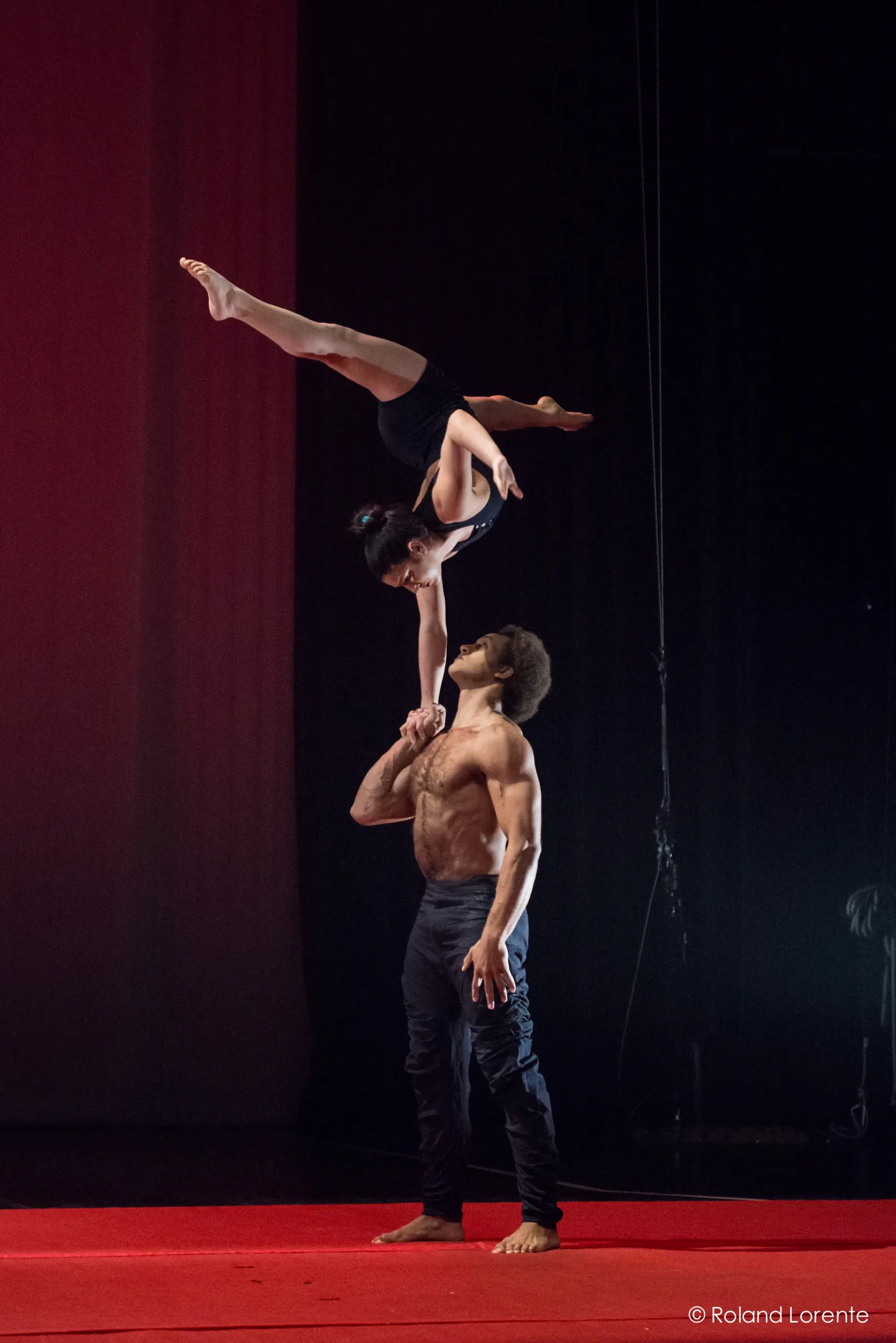 Marlon Archer et Jenay Espinosa se produisent alors qu'un acrobate masculin se tient torse nu sur une scène rouge, faisant tenir sa partenaire à l'envers sur une main au-dessus de sa tête, tous deux concentrés et équilibrés sous l'éclairage de la scène.