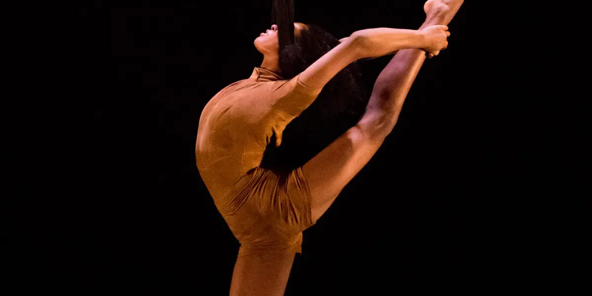 Une artiste aérienne, Tuedon Ariri, vêtue d'un costume marron, étire gracieusement une jambe en l'air tout en se suspendant à une corde aérienne noire sur un fond sombre.