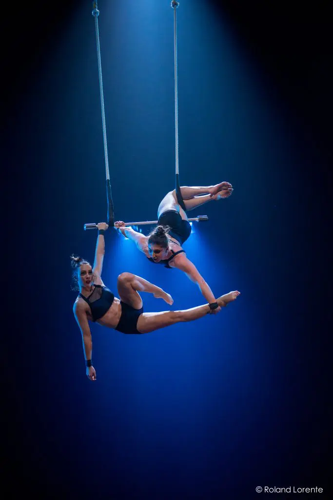 Deux acrobates féminines en tenue noire, dont Virginie Gerbeau, exécutent un numéro aérien sur un trapèze, suspendues dans les airs sous un éclairage de scène bleu spectaculaire. Toutes deux prennent des poses dynamiques, mettant en valeur leur force et leur équilibre.