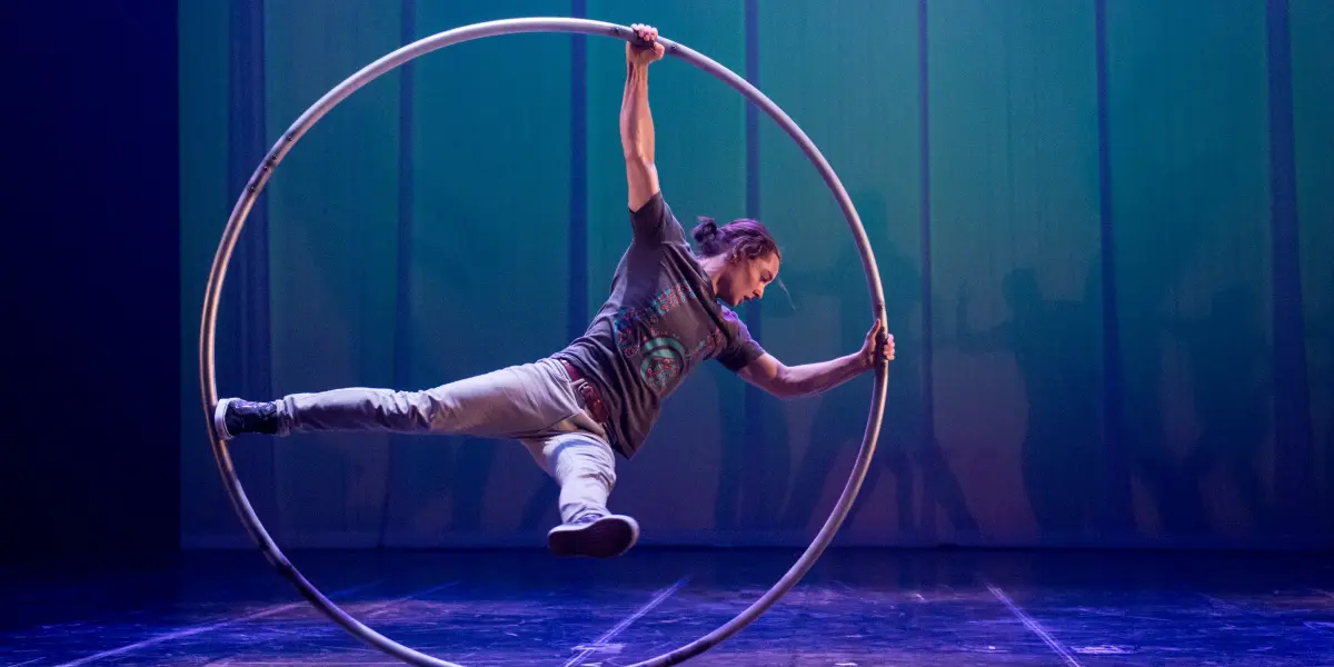 Cory Marsh se tient en équilibre horizontal à l'intérieur d'un grand cerceau métallique sur une scène éclairée en bleu et en vert, tenant le cerceau d'une main et d'un pied tout en étendant l'autre bras et l'autre jambe vers l'extérieur.