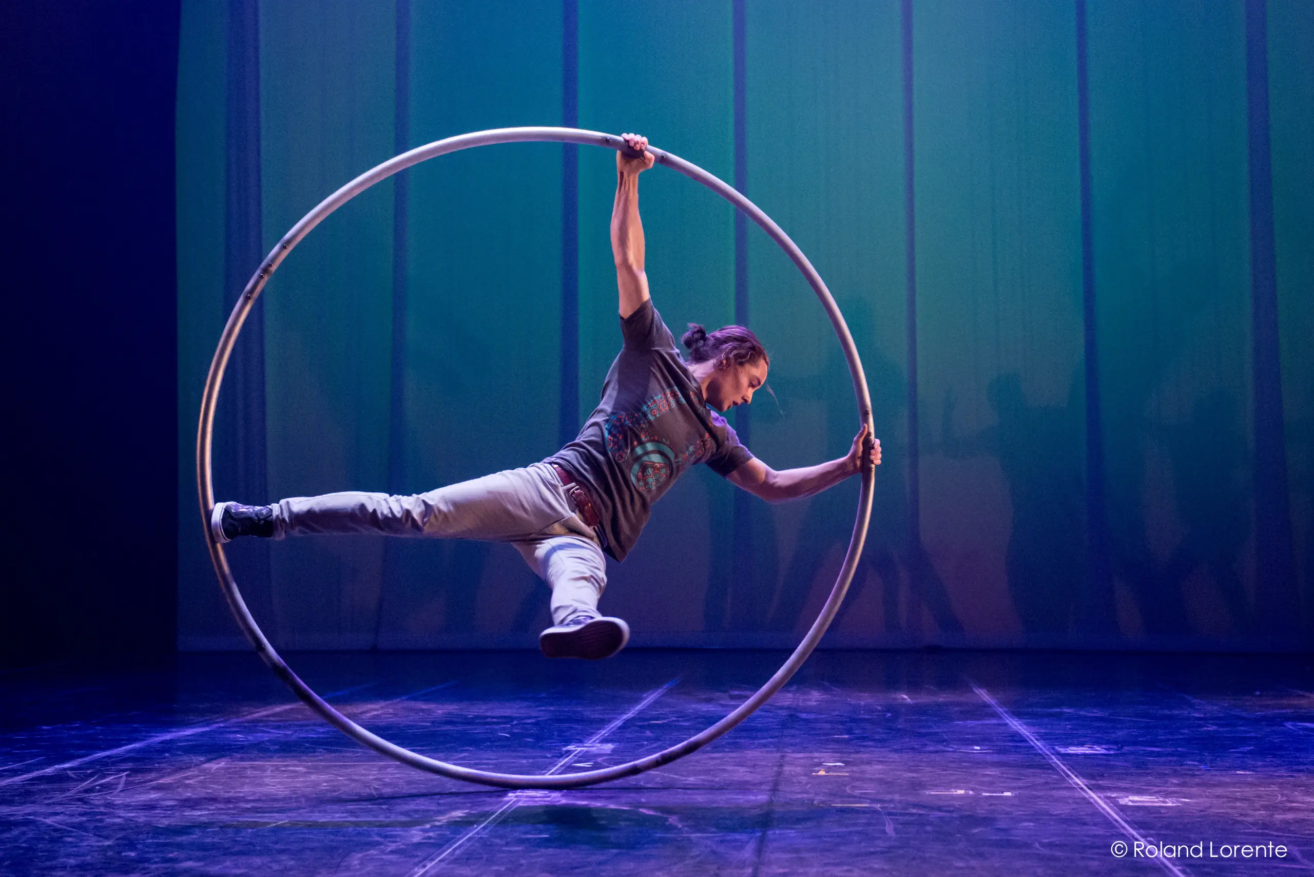 Cory Marsh se tient en équilibre horizontal à l'intérieur d'un grand cerceau métallique sur une scène éclairée en bleu et en vert, tenant le cerceau d'une main et d'un pied tout en étendant l'autre bras et l'autre jambe vers l'extérieur.
