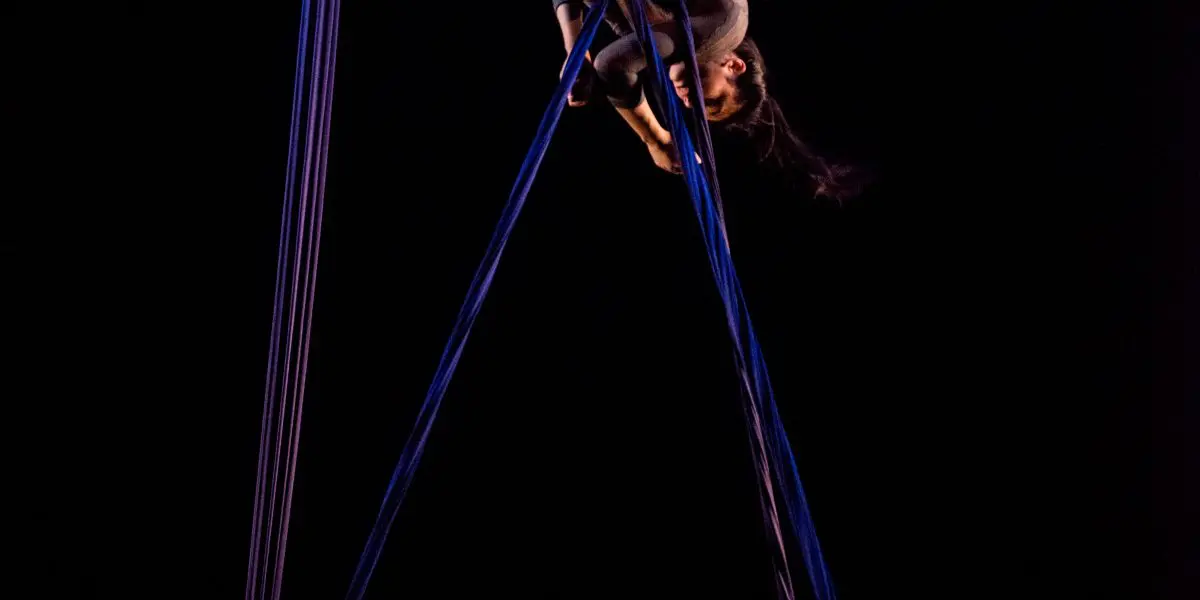 L'artiste aérienne Vanessa Aviles se produit en hauteur, suspendue à l'envers dans une pose dramatique avec des soies bleues, sur un fond sombre.