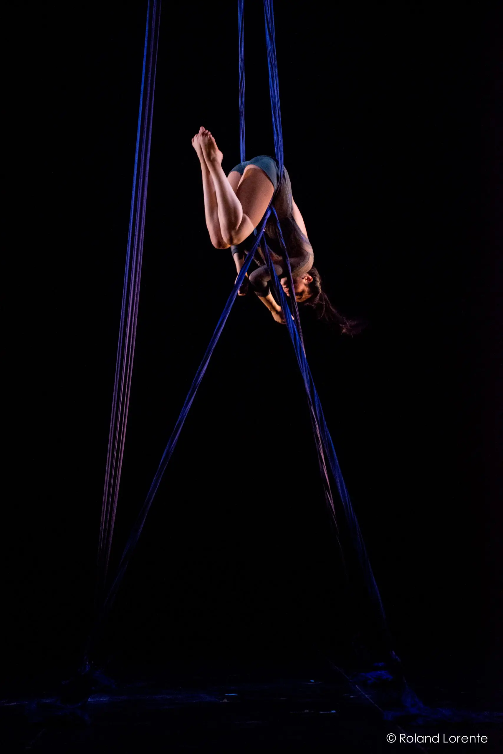 L'artiste aérienne Vanessa Aviles se produit en hauteur, suspendue à l'envers dans une pose dramatique avec des soies bleues, sur un fond sombre.
