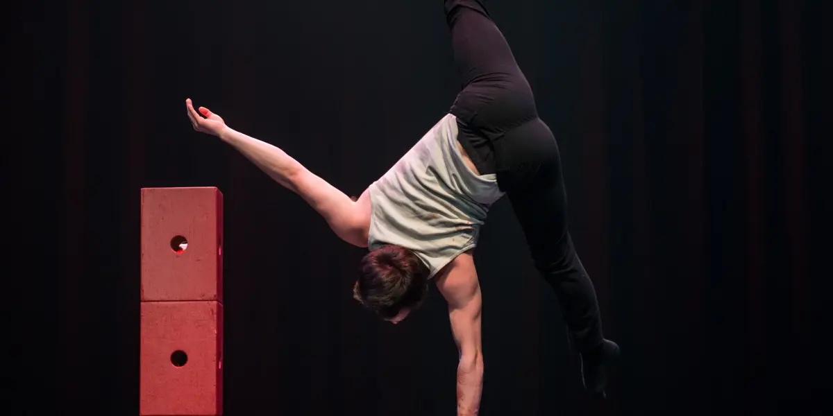 Florian Jeannot, un artiste de cirque expérimenté, se tient en équilibre sur une main au sommet de barres parallèles, avec une jambe tendue vers le haut et l'autre pliée. Des blocs rouges sont empilés à côté de lui sur une scène sombre et éclairée.