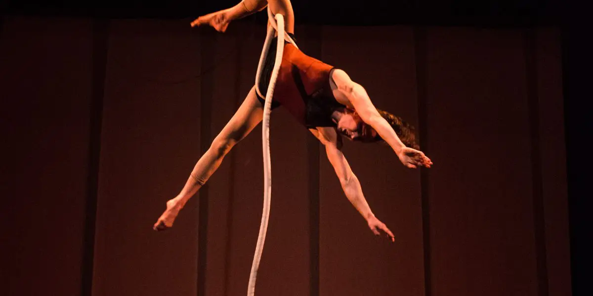 Rosita Hendry, une acrobate aérienne, se produit au-dessus de la scène, suspendue à l'envers à une corde blanche, les bras et les jambes étendus gracieusement dans une pose dramatique sur une toile de fond sombre.